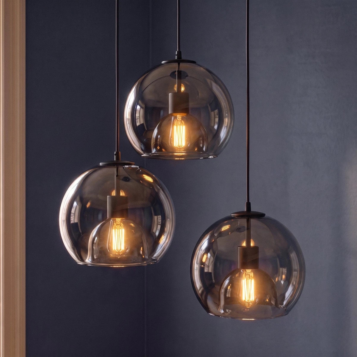 Clérachic trio of smoked glass globe pendants with matte black fittings and amber filament bulbs against a slate blue wall.
