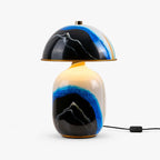This Clérachic table lamp on a white background, with a black dome shade over a ceramic base painted with a blue and black mountain, cord and switch beside it.