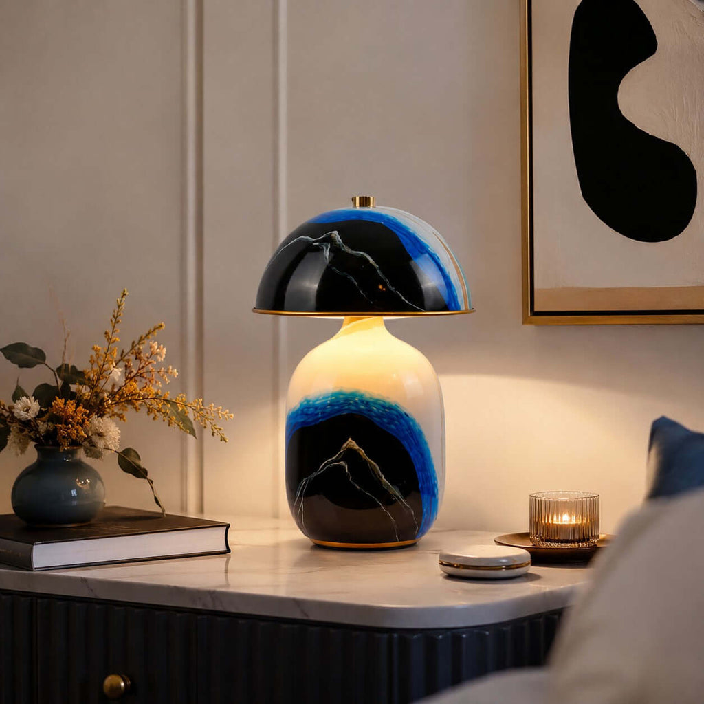 A lit Clérachic table lamp on a fluted console with a candle, a book and a vase of dried flowers beside abstract art.