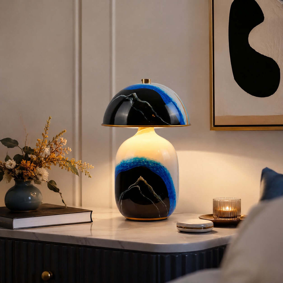 A lit Clérachic table lamp on a fluted console with a candle, a book and a vase of dried flowers beside abstract art.