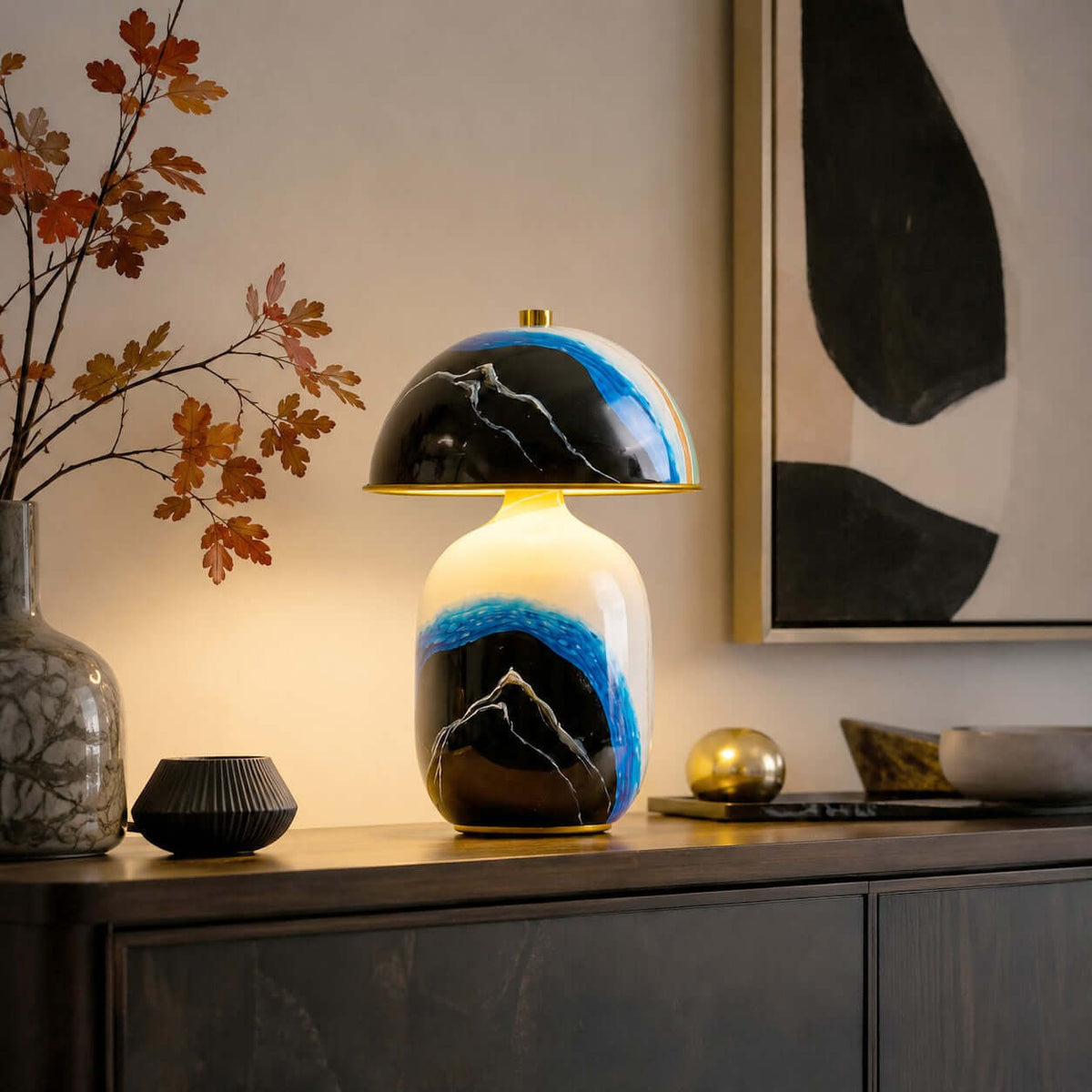Clérachic table lamp glowing on a dark sideboard, with autumn leaves in a vase, a ridged black bowl and a gold sphere nearby.