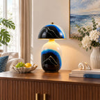 Hand-painted mountain base of this Clérachic table lamp on a wooden sideboard, beside a shell dish of fruit and a seascape painting.