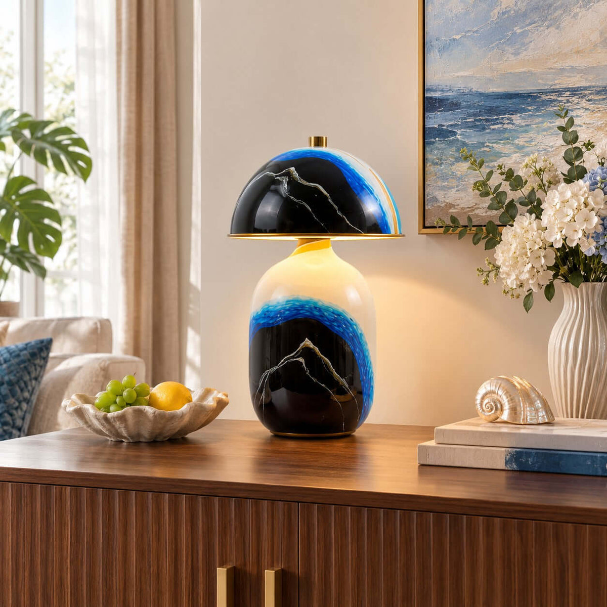Hand-painted mountain base of this Clérachic table lamp on a wooden sideboard, beside a shell dish of fruit and a seascape painting.