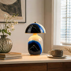 A Clérachic table lamp on a pale console by a sunny window, with a stack of books, a coffee cup and a glass vase of white flowers.