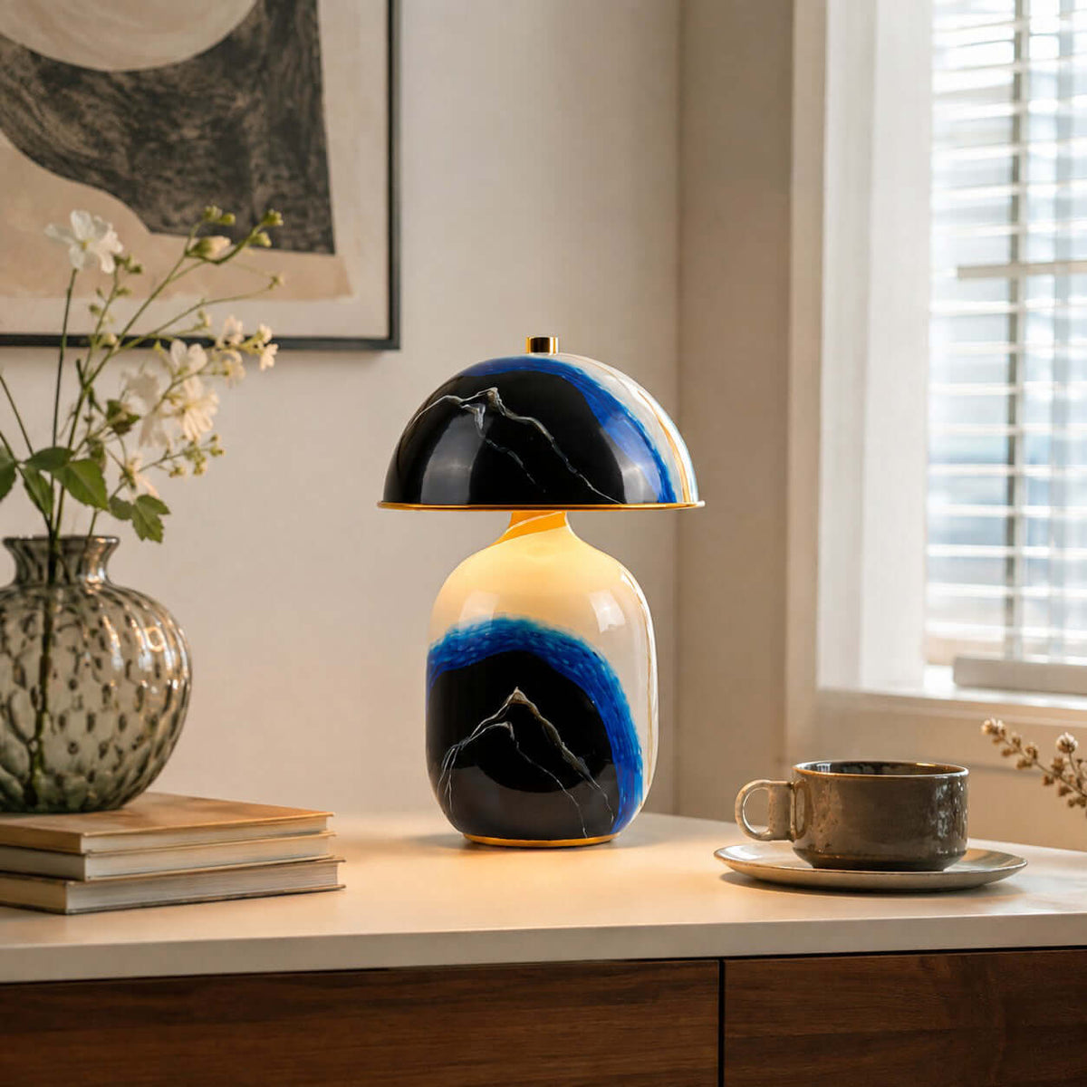 A Clérachic table lamp on a pale console by a sunny window, with a stack of books, a coffee cup and a glass vase of white flowers.