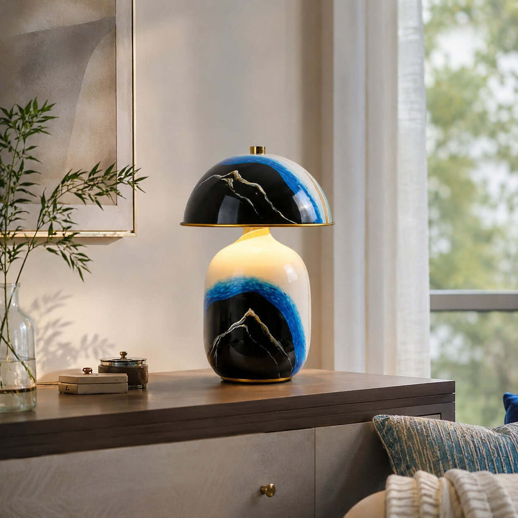 Clérachic table lamp on a dark wooden console near a window, with a leafy branch, small trays and blue cushions at right.