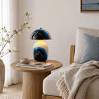 A Clérachic table lamp on a round fluted side table between a linen sofa and a window with white curtains, with books and a bowl.