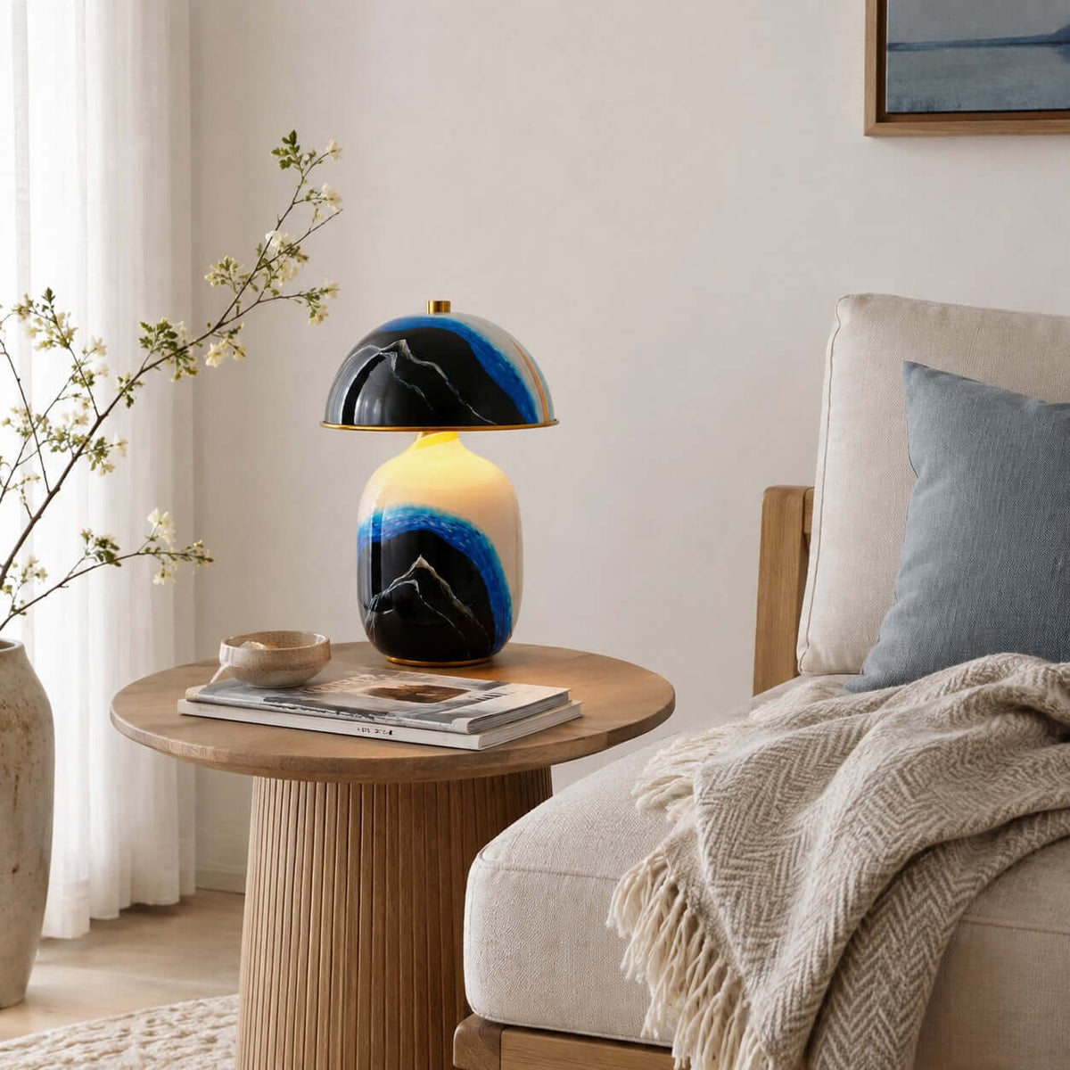 A Clérachic table lamp on a round fluted side table between a linen sofa and a window with white curtains, with books and a bowl.