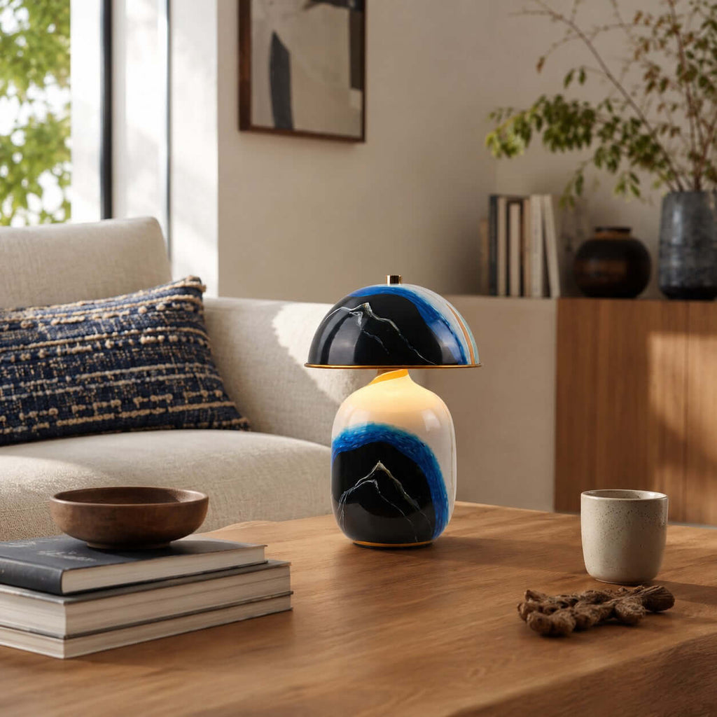 Close view of a Clérachic table lamp on a wooden coffee table with a stack of books, a wooden bowl, a cup and a navy cushioned sofa.