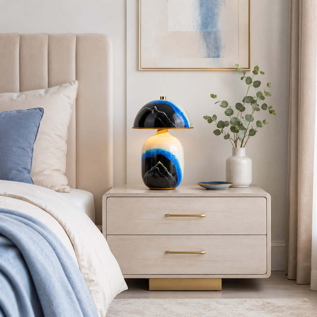 A Clérachic table lamp on a pale two-drawer bedside chest with gold handles, beside a bed with blue bedding and a vase of eucalyptus.