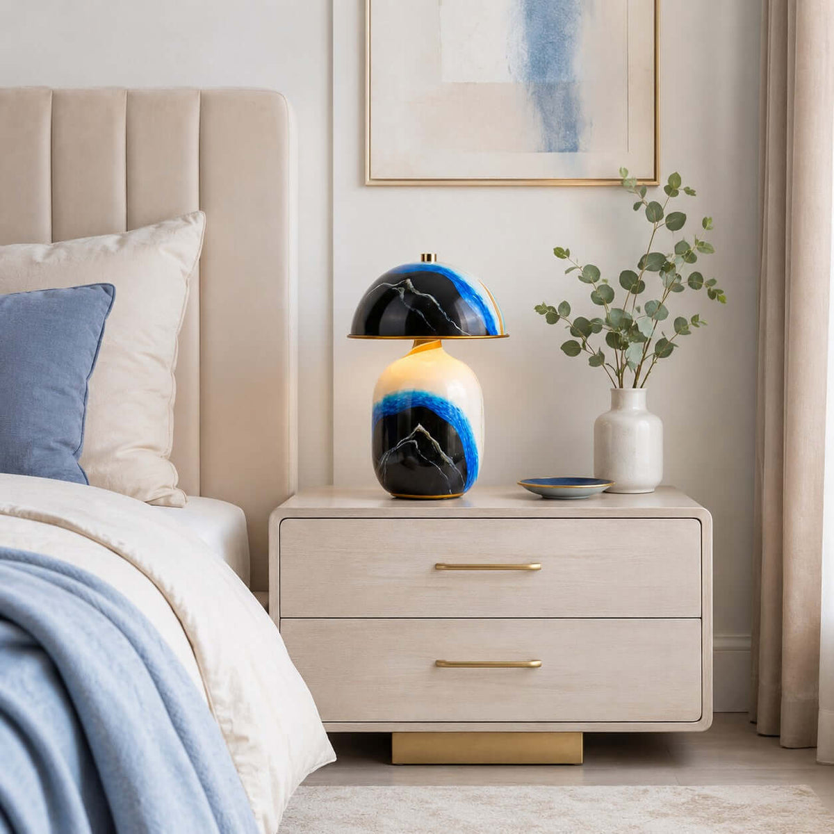 A Clérachic table lamp on a pale two-drawer bedside chest with gold handles, beside a bed with blue bedding and a vase of eucalyptus.