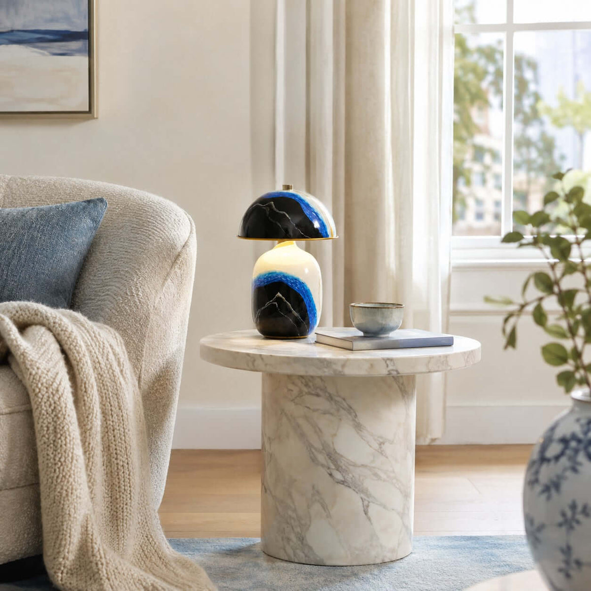 Clérachic table lamp on a round marble side table next to a boucle chair with a cream throw, a bowl and a book.