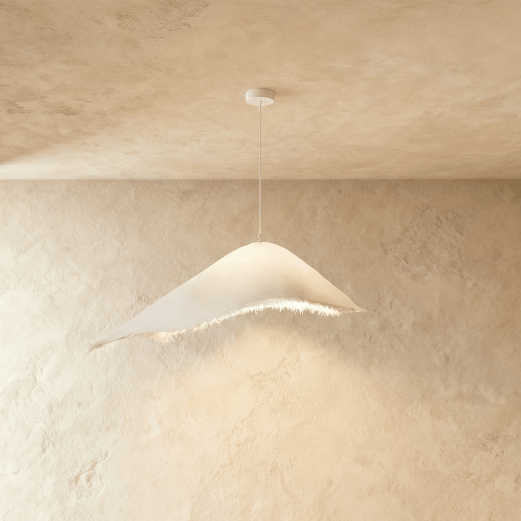 A Clérachic draped white shade swoops low over a fringed edge, hung on a thin cord against a plastered corner.