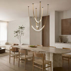 Over a long travertine dining table with oak chairs, this Clérachic pendant sweeps down on brass-toned straps, its lowest arc brightly lit.