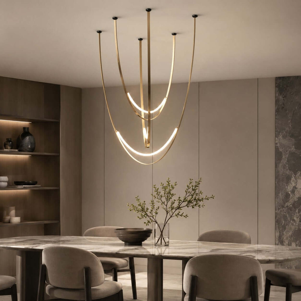 Above a marble table and rounded grey chairs, this Clérachic pendant hangs in brass-toned straps with a bright arc beside a stone wall.