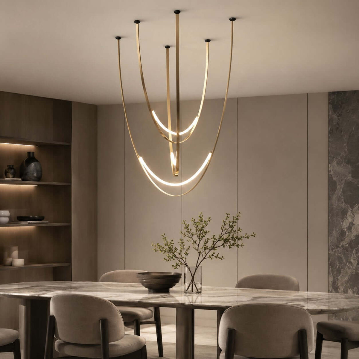 Above a marble table and rounded grey chairs, this Clérachic pendant hangs in brass-toned straps with a bright arc beside a stone wall.