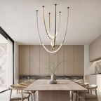 Over a long pale wood table in front of tall veneer cabinets, this Clérachic pendant hangs on brass-toned straps with lit arcs.