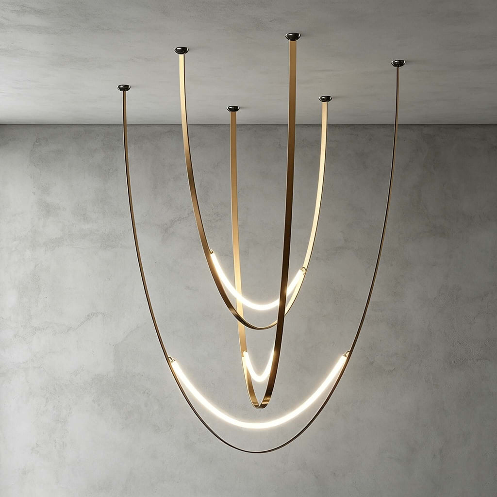 Brass-toned version of this Clérachic pendant against a grey concrete wall, its glowing LED arcs set low on long curving metal straps.