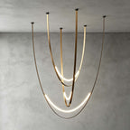 Brass-toned version of this Clérachic pendant against a grey concrete wall, its glowing LED arcs set low on long curving metal straps.