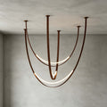 Brown-strap version of this Clérachic pendant against a grey concrete wall, its LED arcs glowing along the long, curved straps.