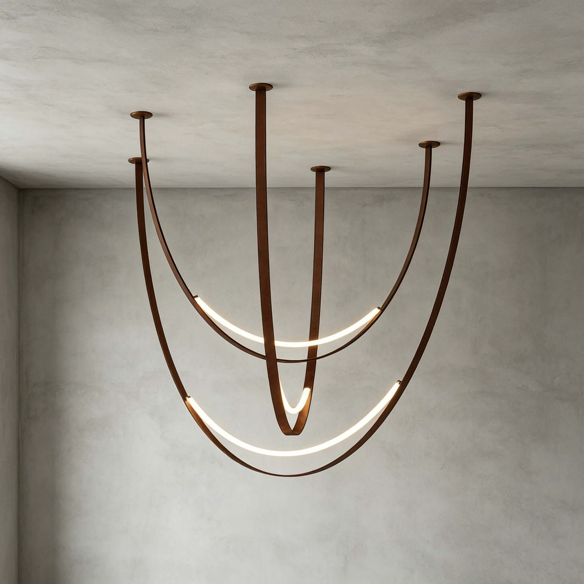 Brown-strap version of this Clérachic pendant against a grey concrete wall, its LED arcs glowing along the long, curved straps.