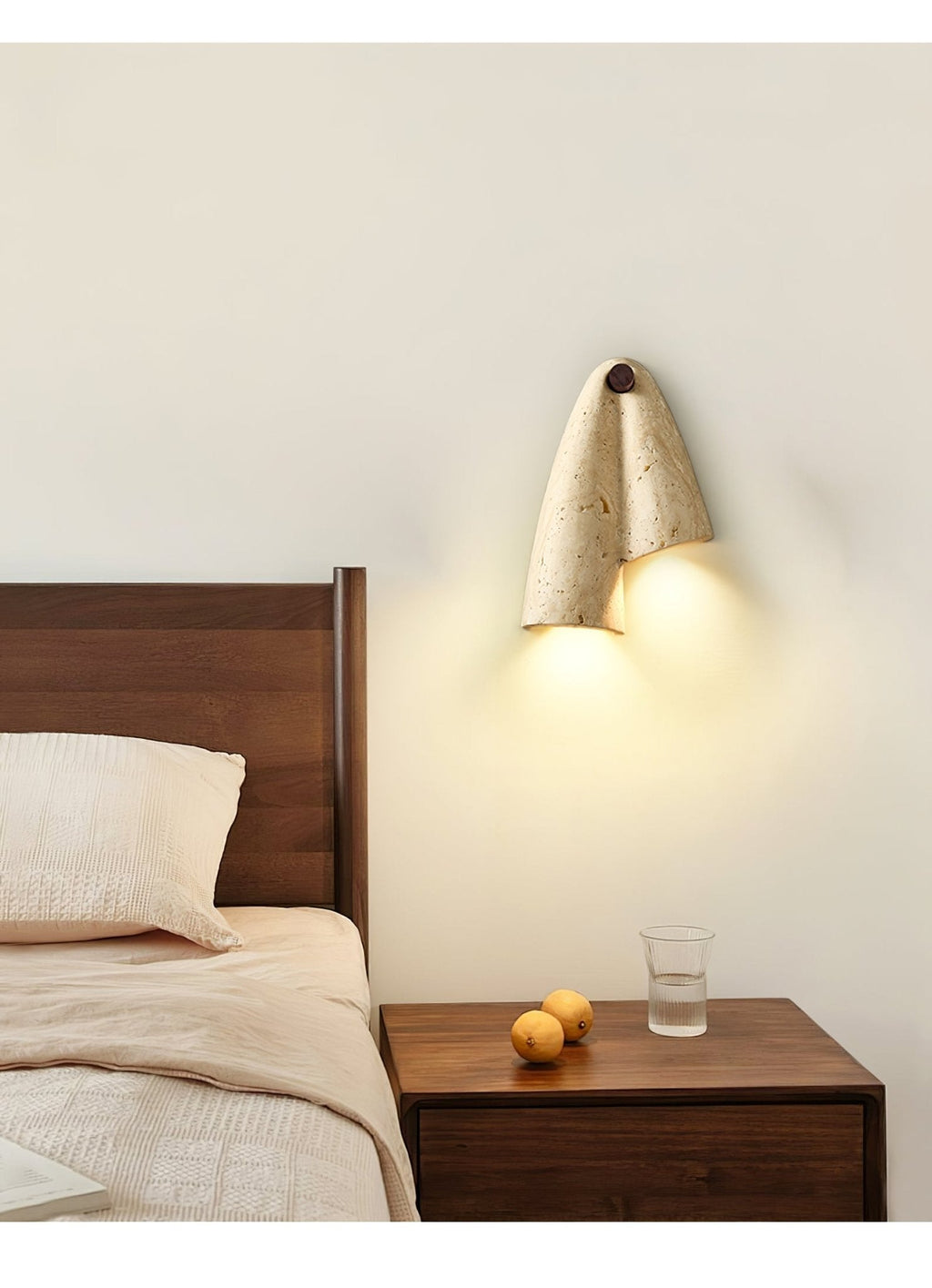 Clérachic travertine wall light above a walnut bedside table, casting a warm glow downward beside a bed with cream linen.