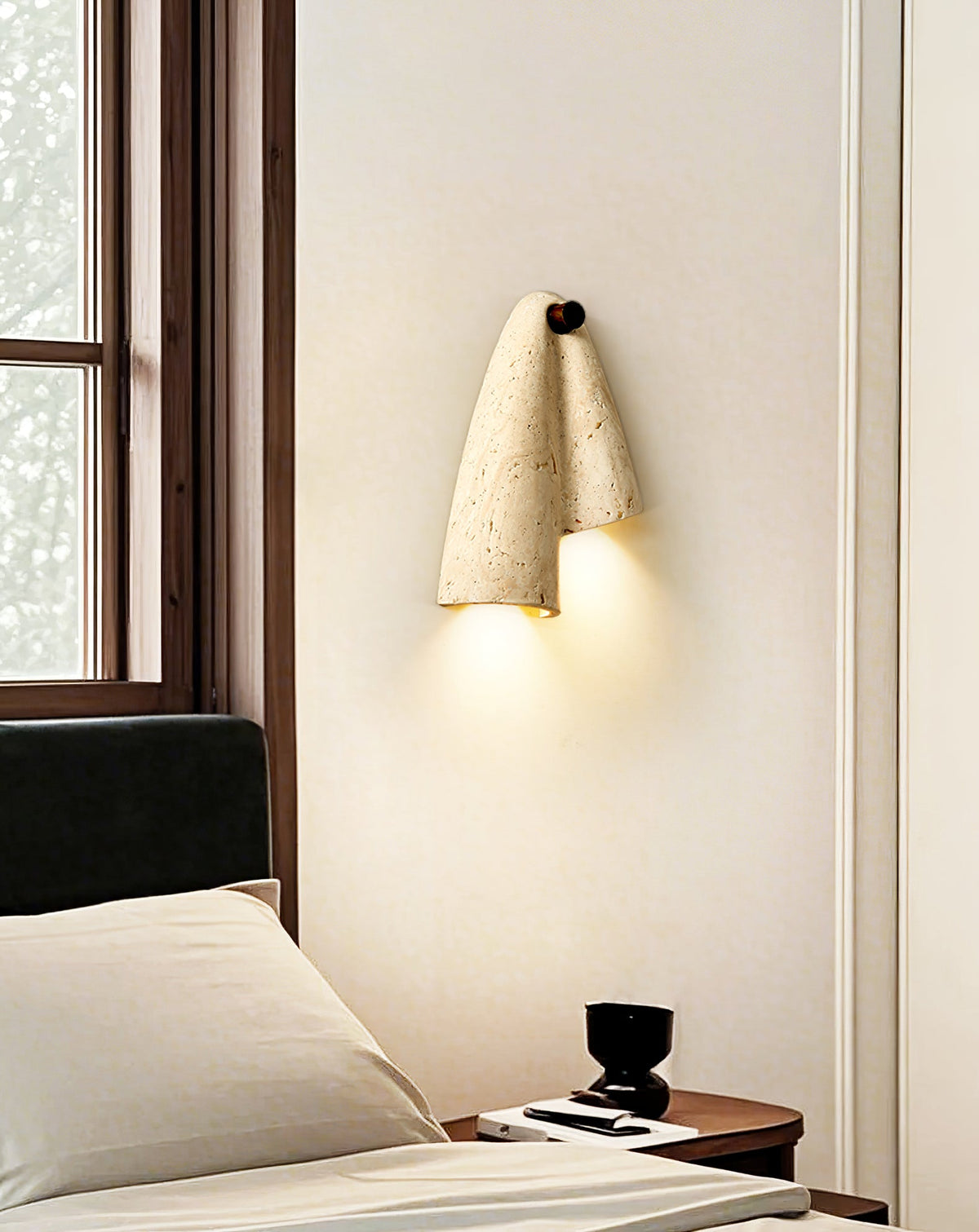 The Clérachic stone sconce lit on a cream wall next to a dark-framed window, above a bed with pale pillows.