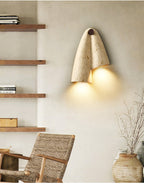 Clérachic draped stone wall light glowing between open wooden shelves, with a woven chair and a clay vase of twigs below.