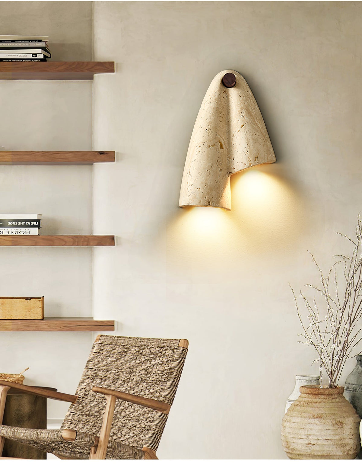Clérachic draped stone wall light glowing between open wooden shelves, with a woven chair and a clay vase of twigs below.