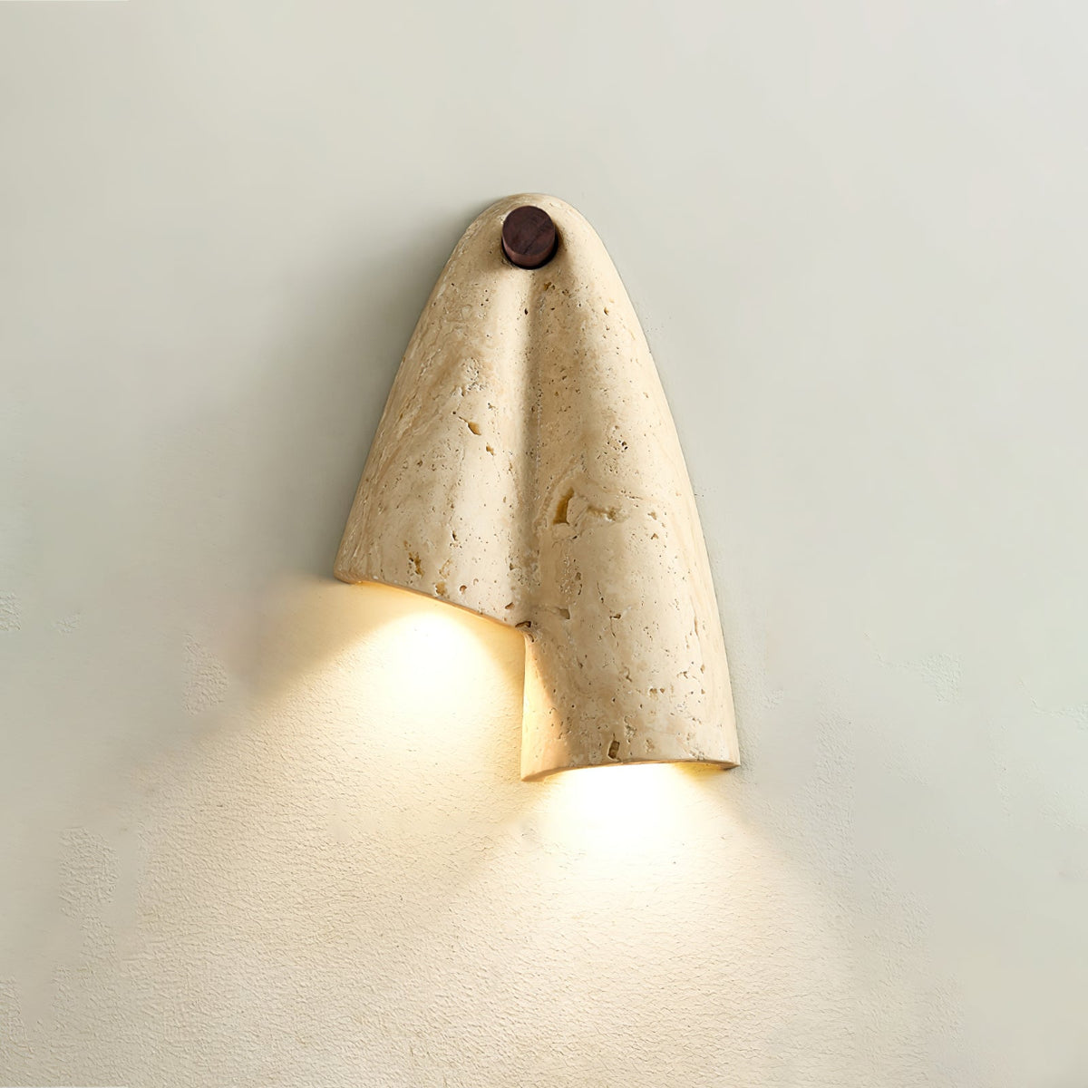 Lit Clérachic travertine wall light on a pale green-grey wall, the fold of stone sending warm light down in two pools.