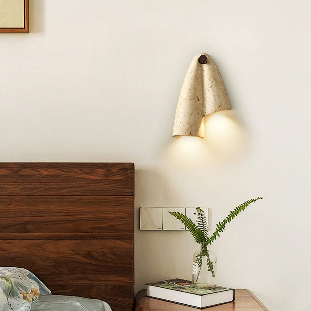 A lit Clérachic wall sconce above a walnut headboard, with a fern in a glass vase and a small stack of books beside it.
