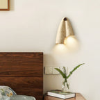 A lit Clérachic wall sconce above a walnut headboard, with a fern in a glass vase and a small stack of books beside it.