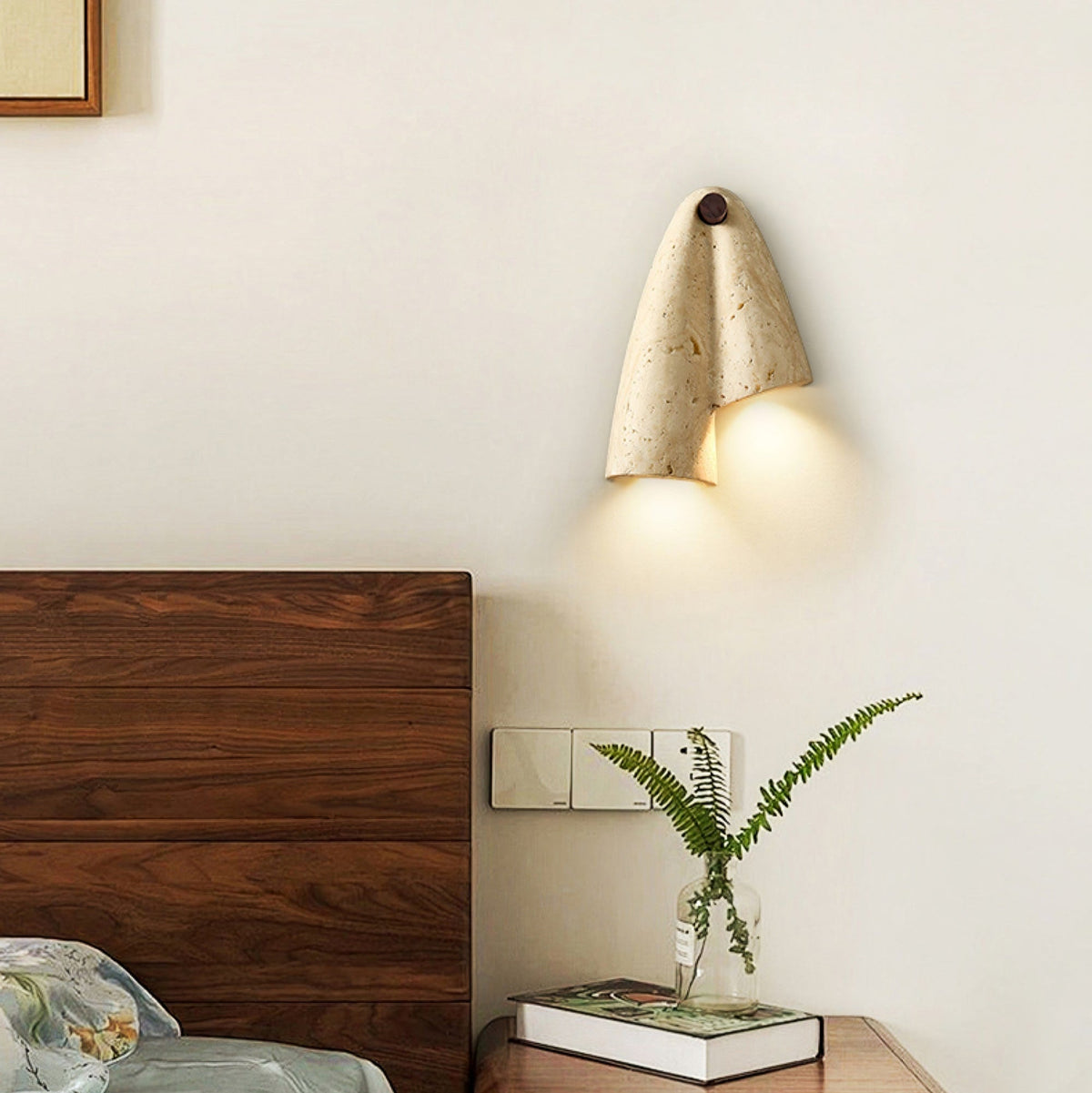 A lit Clérachic wall sconce above a walnut headboard, with a fern in a glass vase and a small stack of books beside it.