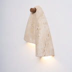 Clérachic travertine wall sconce lit from beneath the fold, with a glow on the wall under the wood knob.