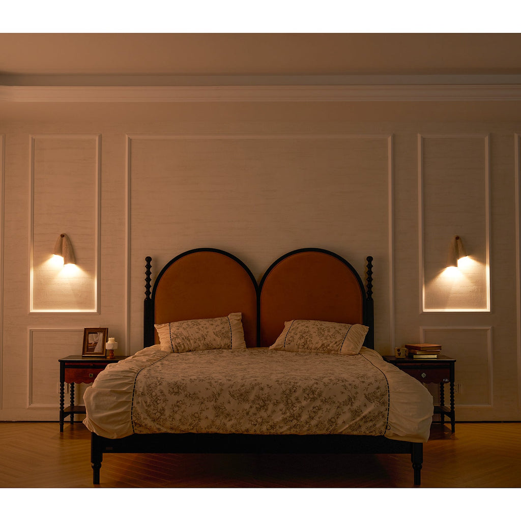 Warm evening bedroom with a Clérachic wall light glowing on each side of a double bed with arched, rust-coloured headboards.