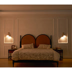 Warm evening bedroom with a Clérachic wall light glowing on each side of a double bed with arched, rust-coloured headboards.