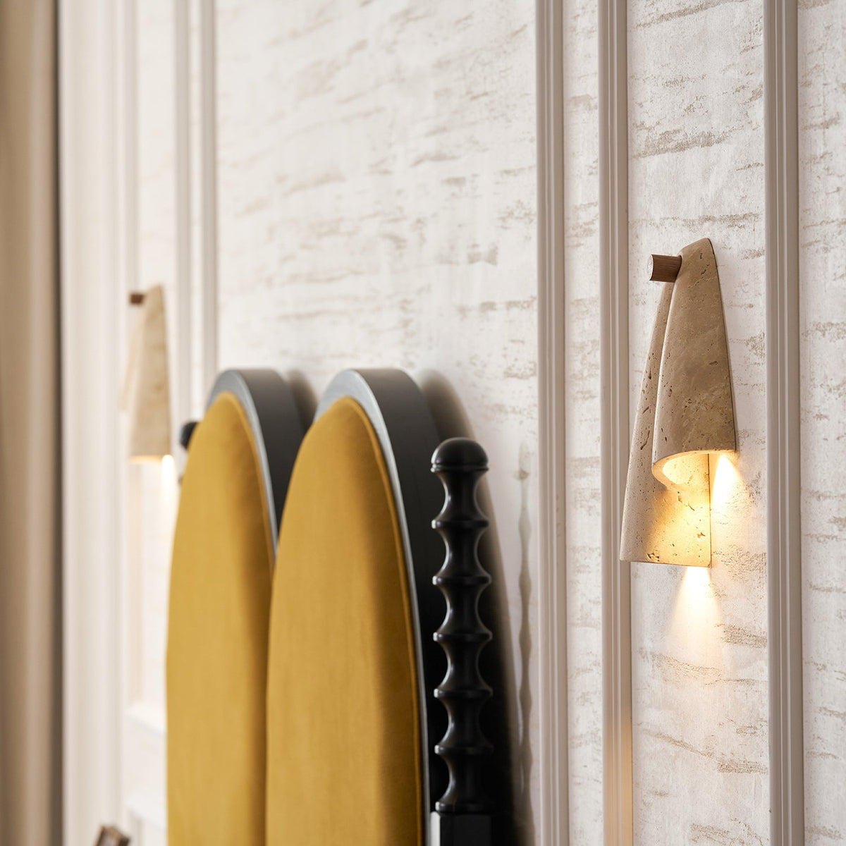 A Clérachic stone wall light glowing on a travertine-look wall, with the edge of a mustard headboard and black turned posts.