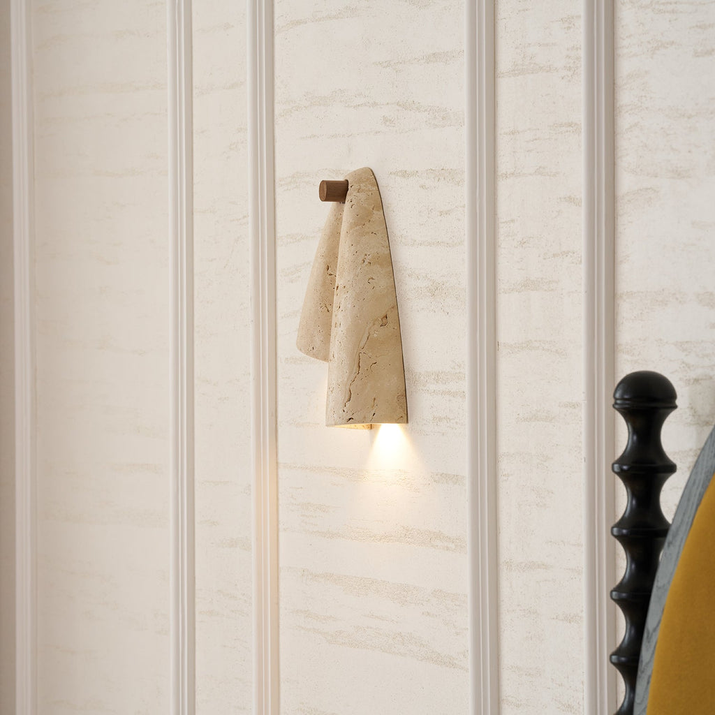 The Clérachic draped sconce lit on a panelled cream wall, with a black bedpost at the right edge.