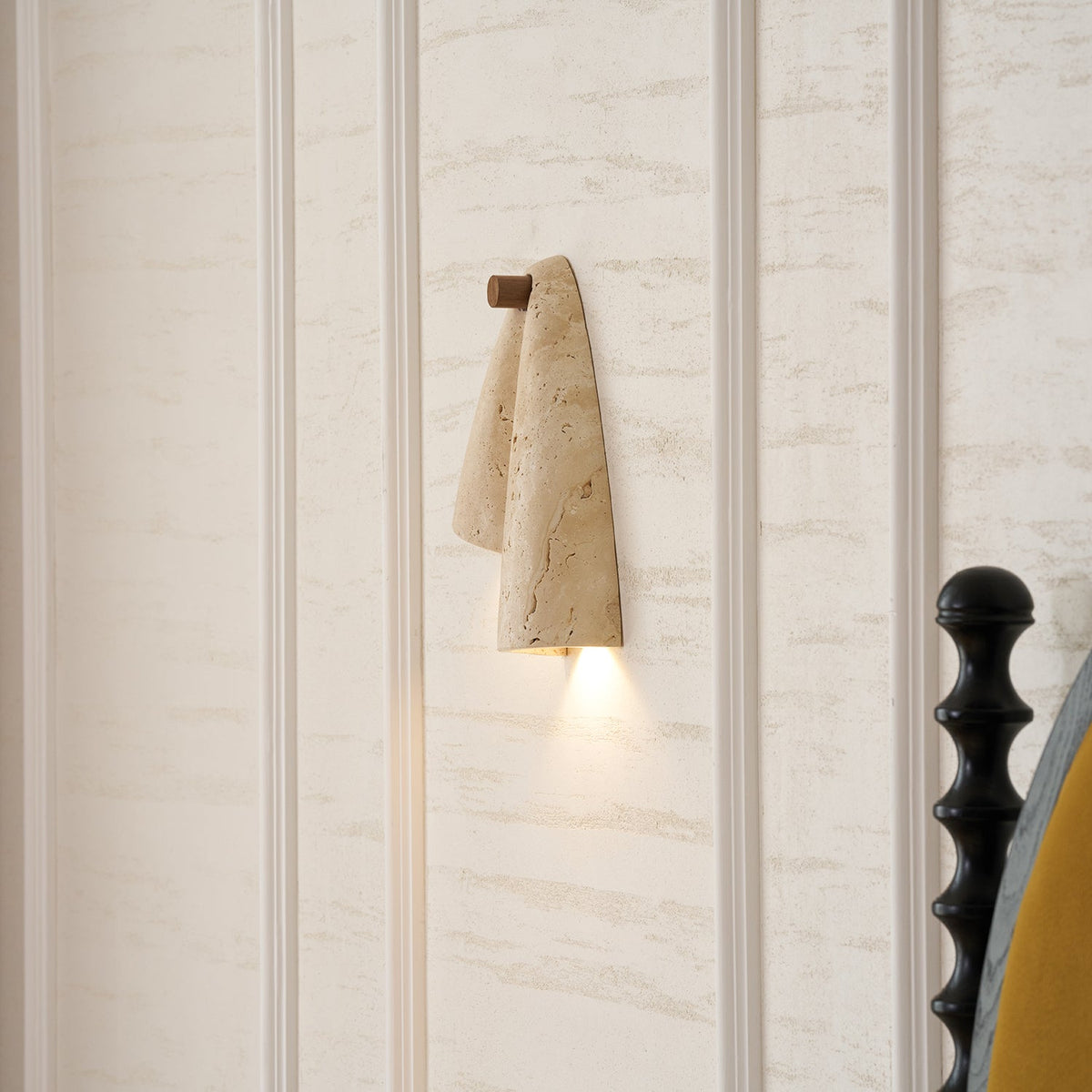 The Clérachic draped sconce lit on a panelled cream wall, with a black bedpost at the right edge.