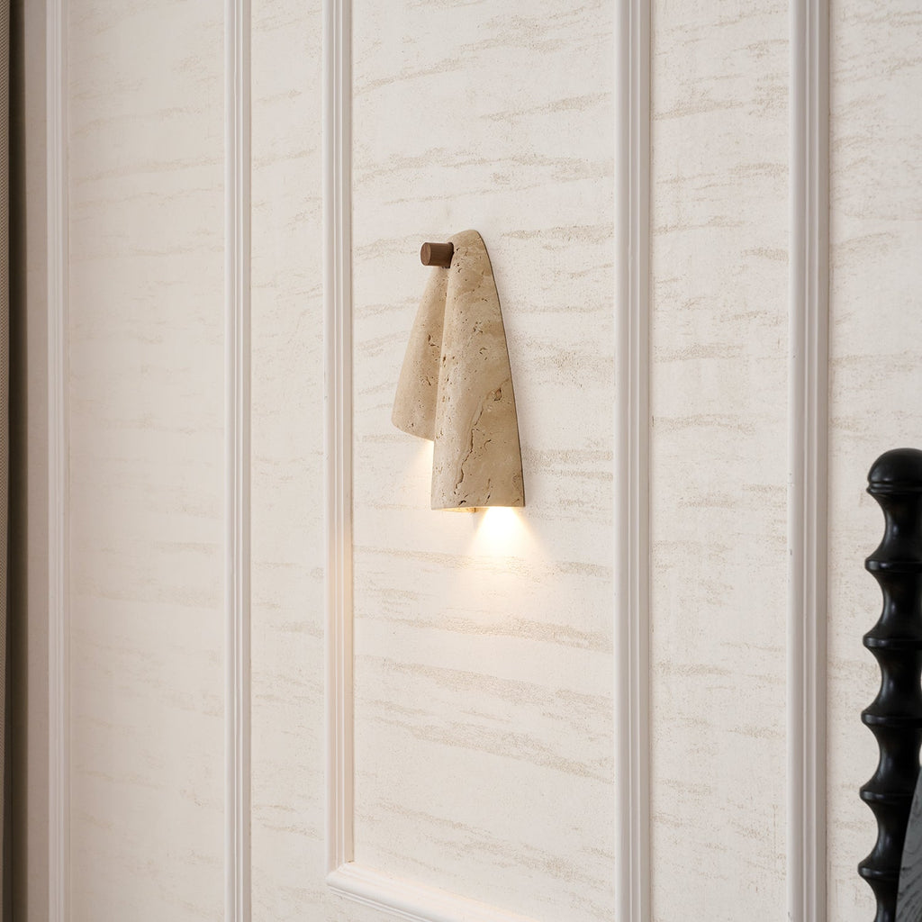 Clérachic stone wall light glowing against a travertine-effect wall with moulded panels and a black bedpost nearby.