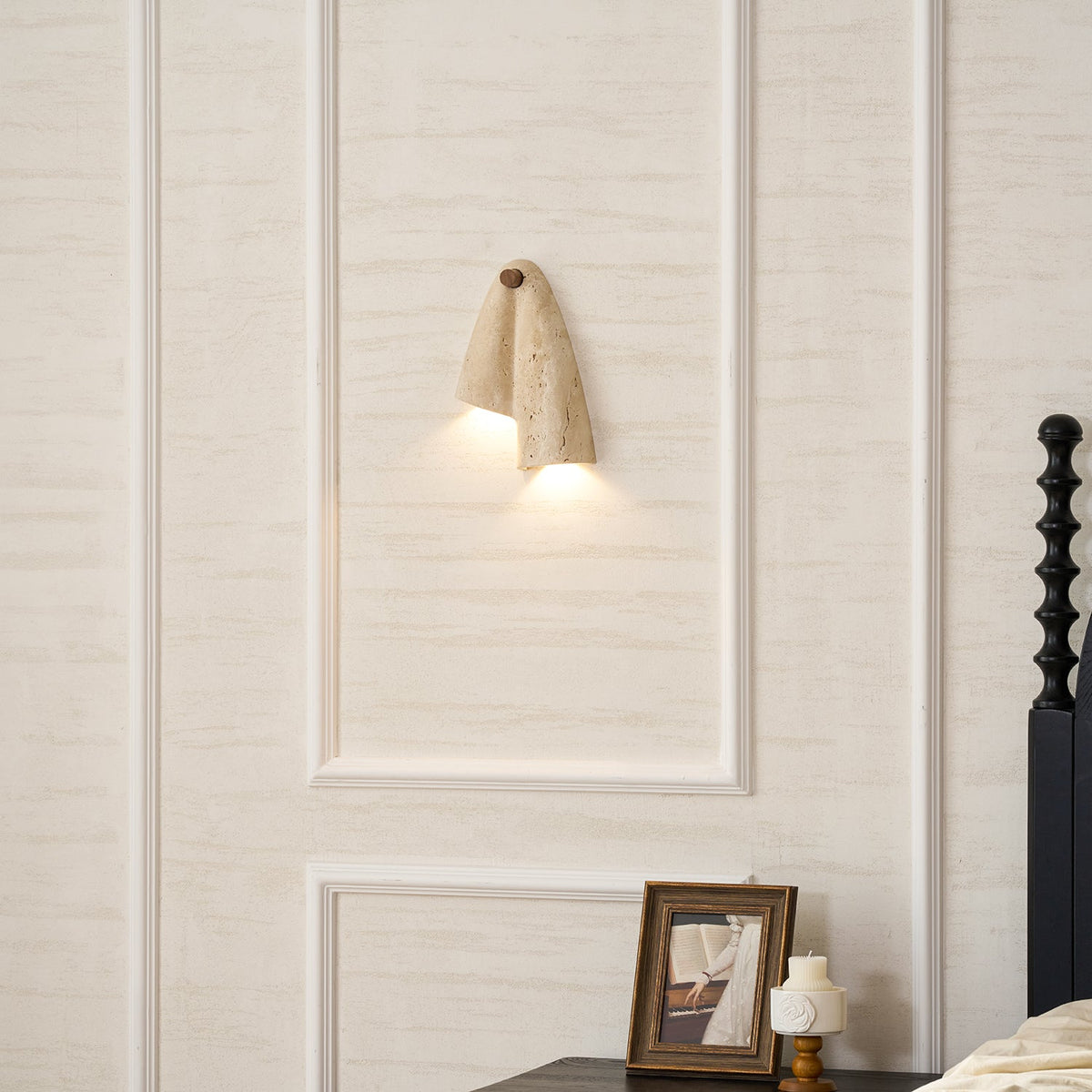 Clérachic sconce lit on a panelled wall above a framed photograph on a bedside table, seen from further back.