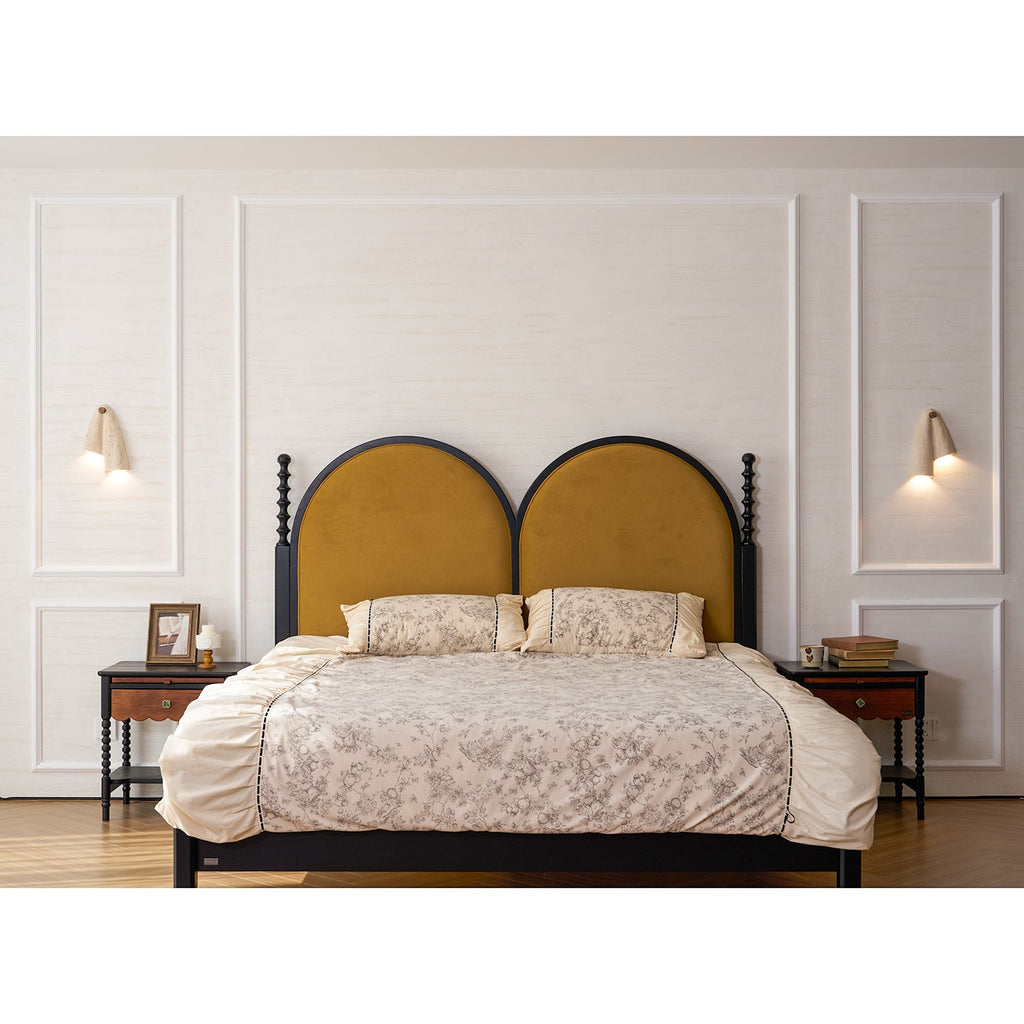 Two Clérachic wall lights flanking a bed with an arched mustard headboard, floral bedding and wood bedside tables.