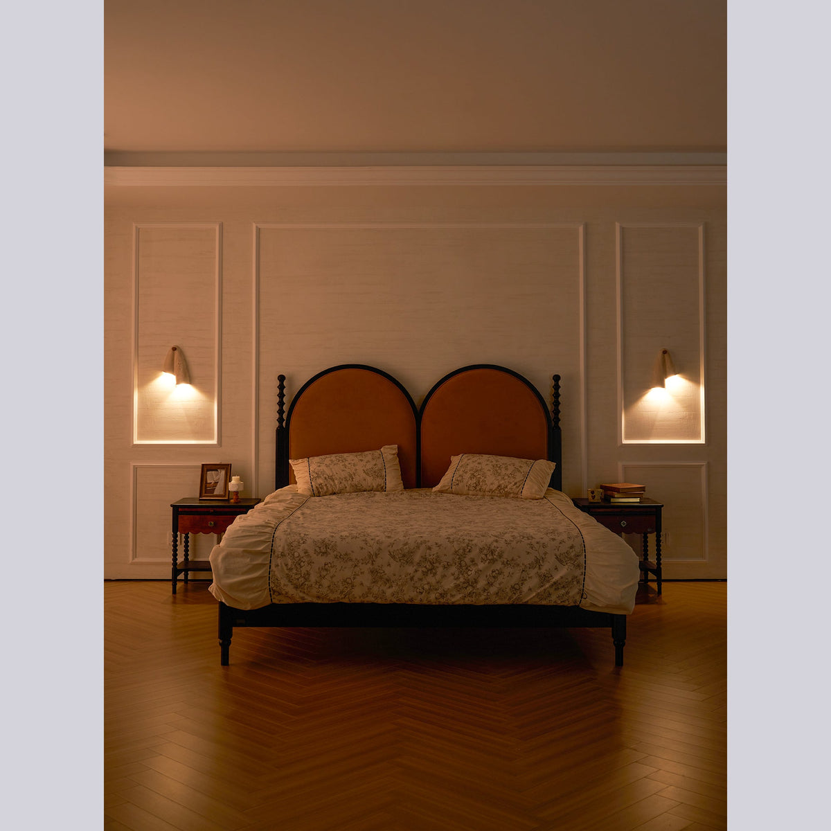 Evening bedroom with a Clérachic wall light on each side of an arched headboard, the room lit warm and the floor in herringbone wood.