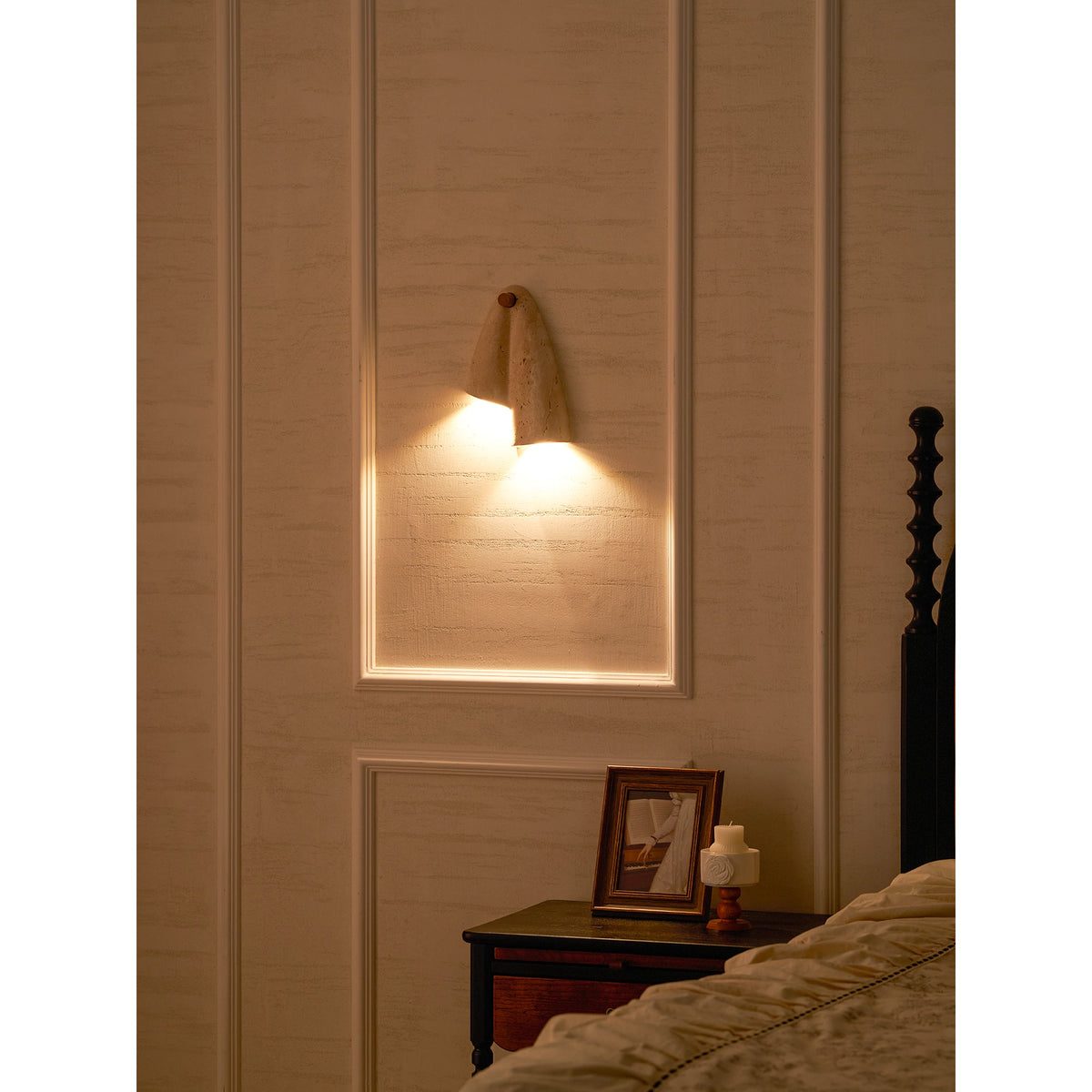 Clérachic stone sconce glowing in a lit wall panel above a bedside table with a framed photo and a small candle.