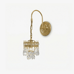 Gold-toned arched wall sconce with a round embossed backplate and a small crystal-drop shade, on a plain white backdrop, from Clérachic.