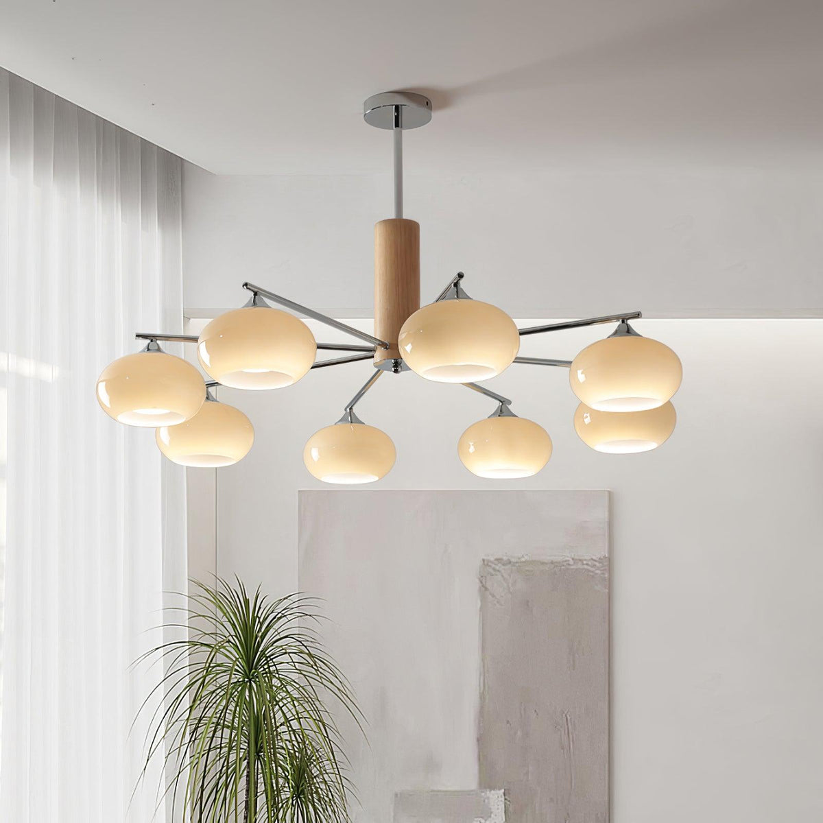 Lit chandelier from Clérachic with chrome arms and creamy glass shades hung in a bright living room, with a tall grass plant and soft curtains beside it.