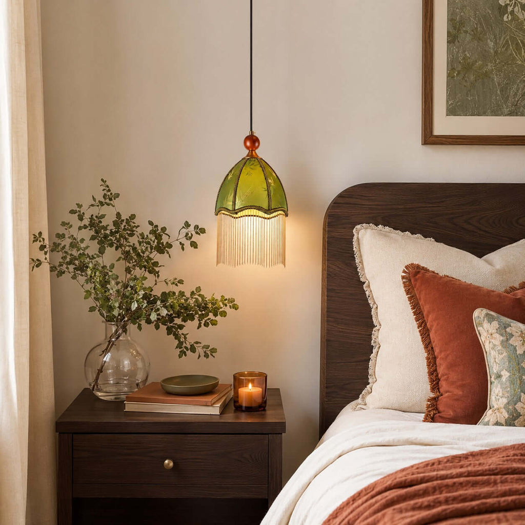 A glowing Clérachic pendant with an olive fabric shade and bead fringe hangs beside a dark wood bedside table and a lit candle.