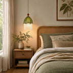 A warm-lit Clérachic pendant with a green fabric dome and fringe hangs beside a rattan headboard and a sage-green bedspread.
