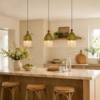 Three Clérachic pendants with scalloped green shades and fringe hang above a kitchen island with rattan stools.