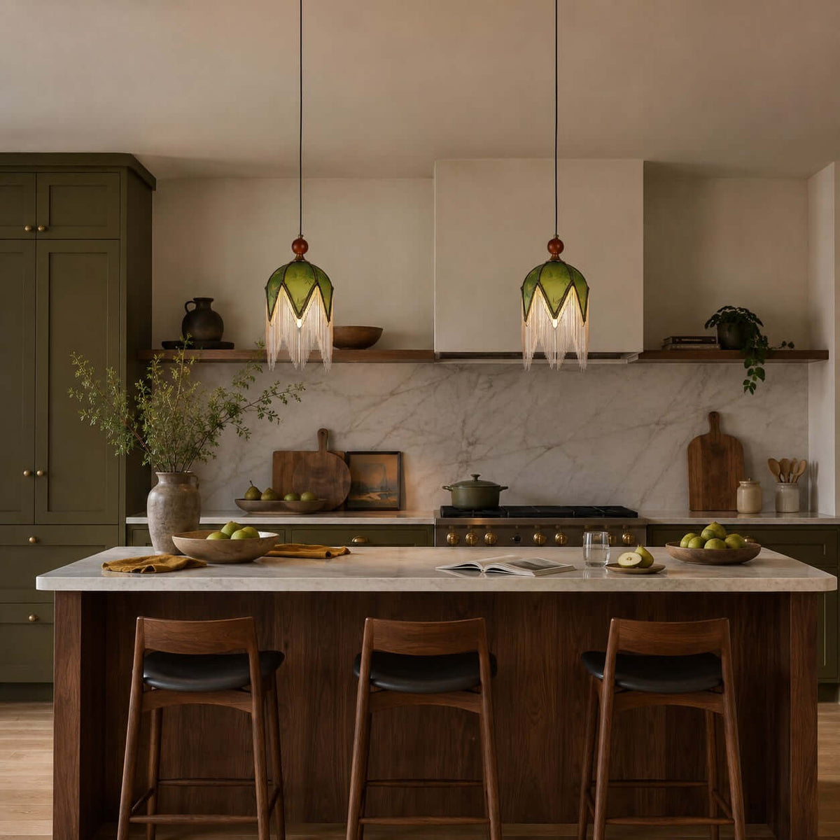 Two Clérachic pendants with green shades and short fringe hang in a dark green kitchen above a walnut island with leather stools.